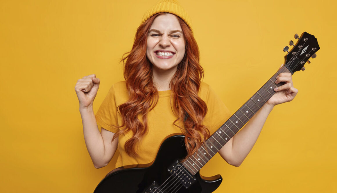 Upbeat redhead woman clenches fist with triumph glad to have many fans plays rock music on electric guitar wears hat and t shirt isolated over yellow background feels cool. Cheerful musician