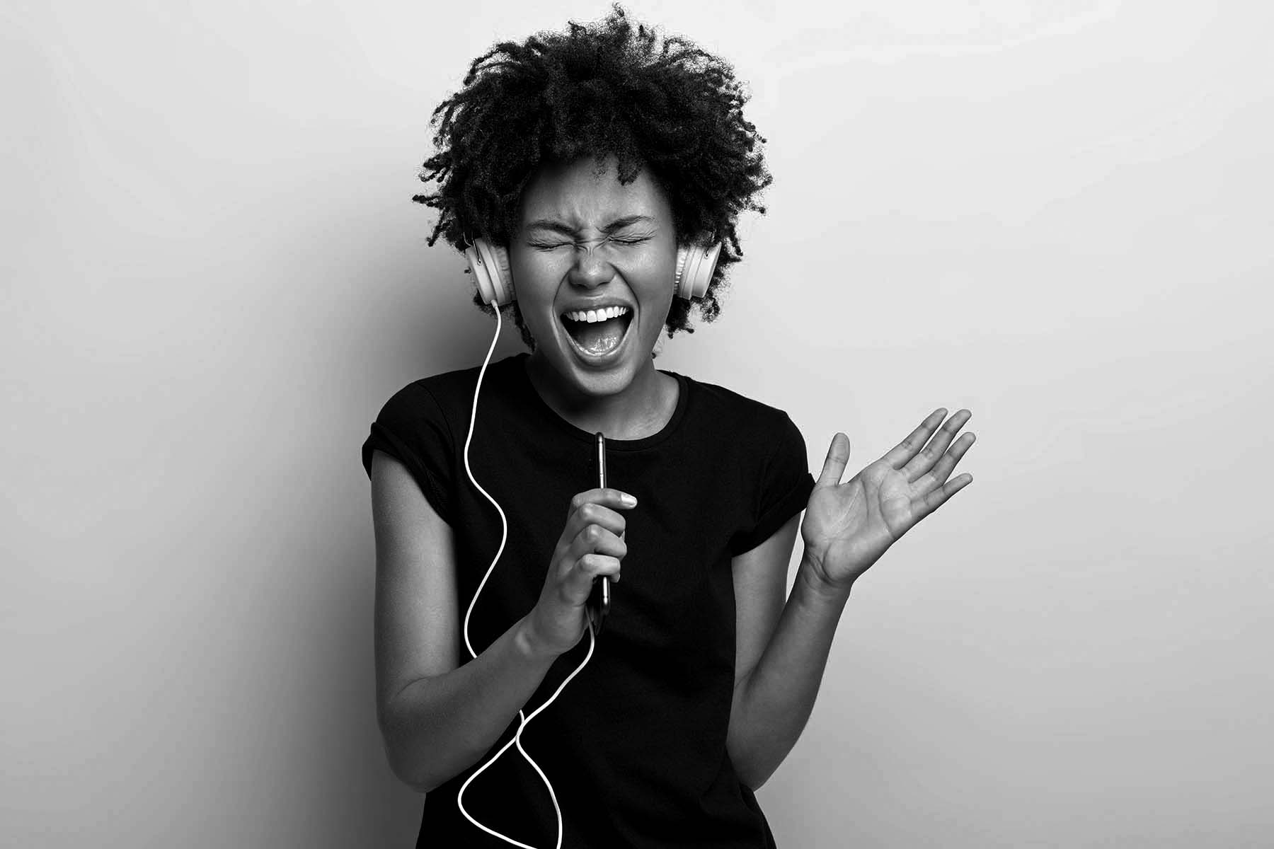 Amused black young woman with curly hair, shakes hands, enjoys cool music, holds smart phone, perfect sound in headphones, has heartbreaking moment, listens favourite track, isolated on yellow